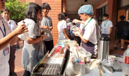 Students at a bake sale stall outdoors on campus; one student serves desserts while others line up to purchase items, surrounded by baked goods, utensils, and cooking supplies, as part of a fundraising event for service learning projects.
